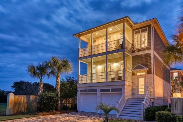 Three-story coastal home with two garages, illuminated wraparound balconies, and palm trees at dusk.