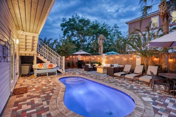 Backyard pool area with a curved blue pool, brick-paver patio, and wicker lounge chairs under string lights and umbrellas