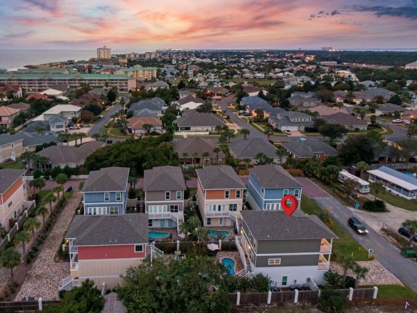 Aerial view of a coastal suburban neighborhood at sunset; row of pastel two‑story homes with backyards and pools, a red pin marks one house in the foreground.