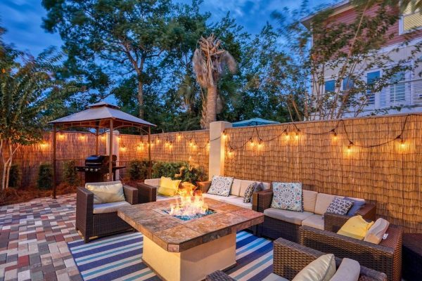 Backyard patio scene with a square fire pit, wicker sectional seating, and string lights along a wooden privacy fence at dusk/evening.
