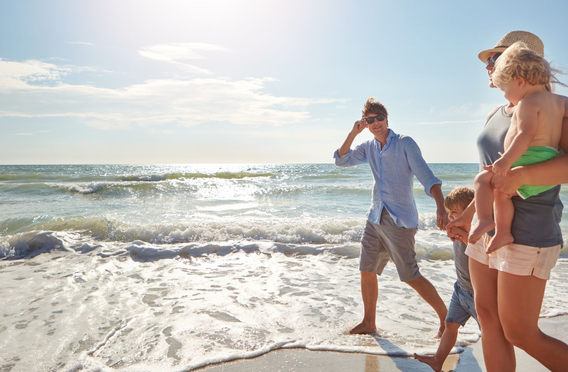 Sunlit beach scene with a family walking along the shore, waves lapping at their feet.