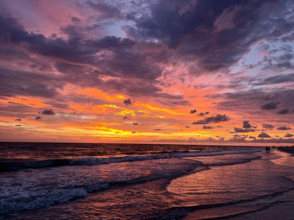 Sunset over the ocean with orange, pink, and purple clouds; waves wash onto a sandy beach where a few people walk in the distance.